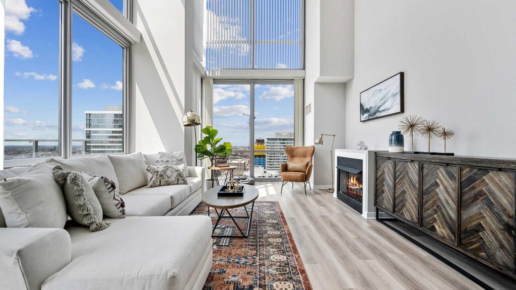 Modern living room with large windows, a white sofa, accent chair, patterned rug, fireplace, and city views visible through the glass.