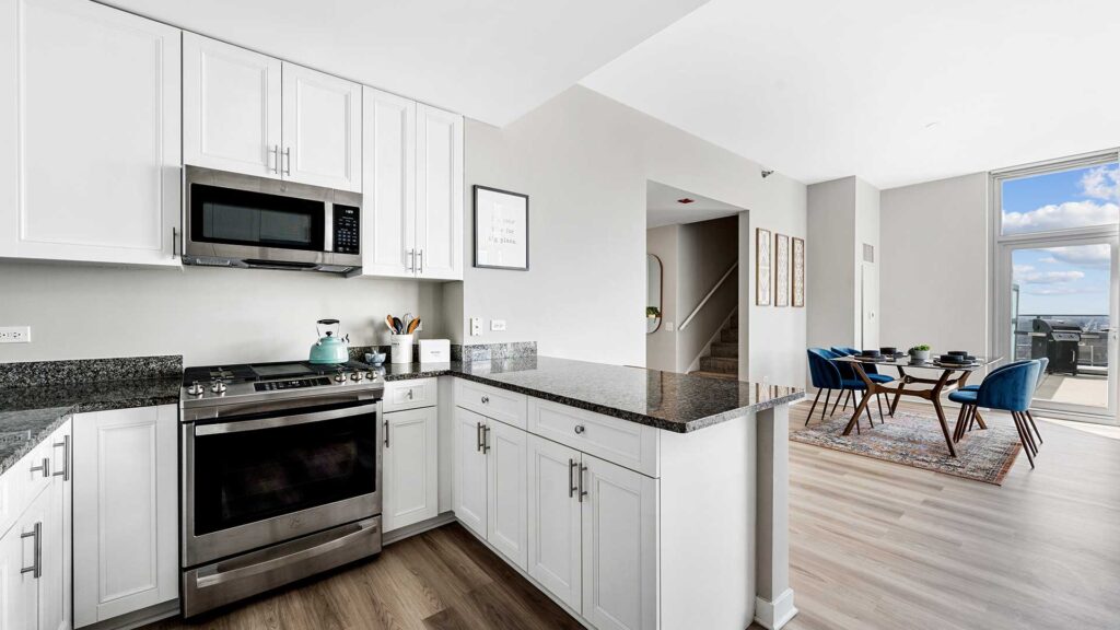 Modern kitchen with white cabinets, stainless steel appliances, granite countertops, and an open layout leading to a dining area with large windows and a view outdoors.
