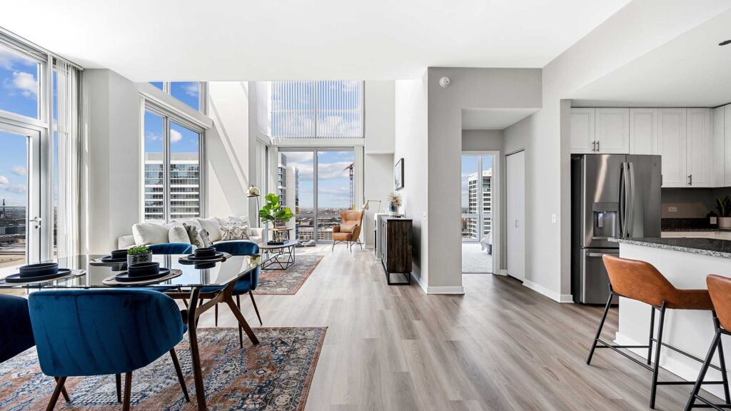 Modern open-concept apartment with large windows, dining area, living room, kitchen with island, and city views. Blue and brown accent furniture, light wood flooring, and natural light.