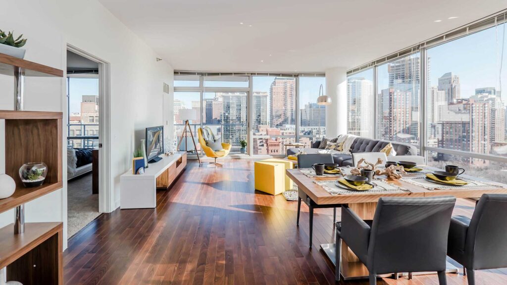 Modern open-concept apartment with floor-to-ceiling windows, wooden floors, dining area, living space, and city skyline views. Bedroom partially visible on the left.
