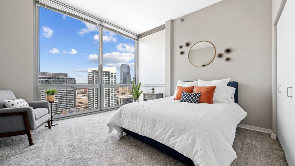 Modern bedroom with large windows showing a cityscape, a neatly made bed with pillows, an armchair, and minimalistic decor.