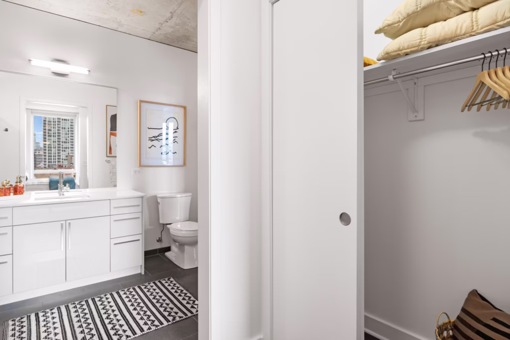 Bathroom with white vanity, toilet, patterned rug, and wall art, next to an open closet with shelves, pillows, hangers, and a small brown bag; city view visible through a window.