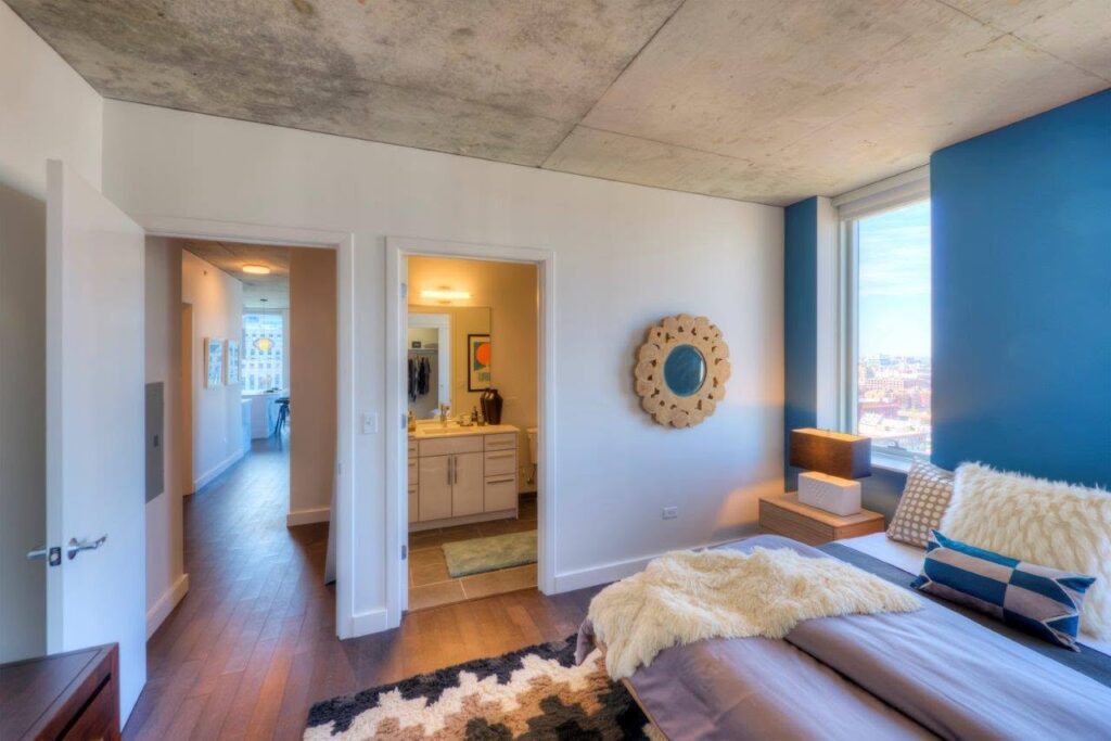 Bedroom with blue accent wall, large window, and modern decor; open doorways lead to a bathroom and hallway with wooden floors and natural light.