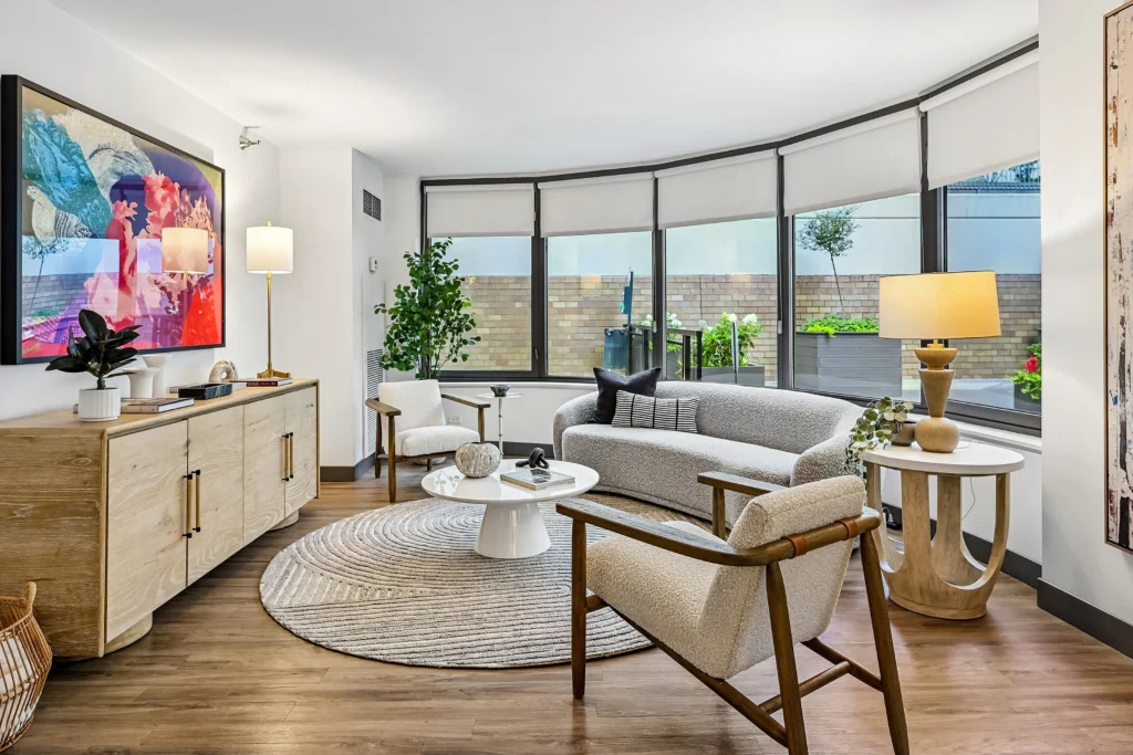 Modern living room with light wood furniture, a round rug, abstract wall art, and large windows showing a patio with plants.