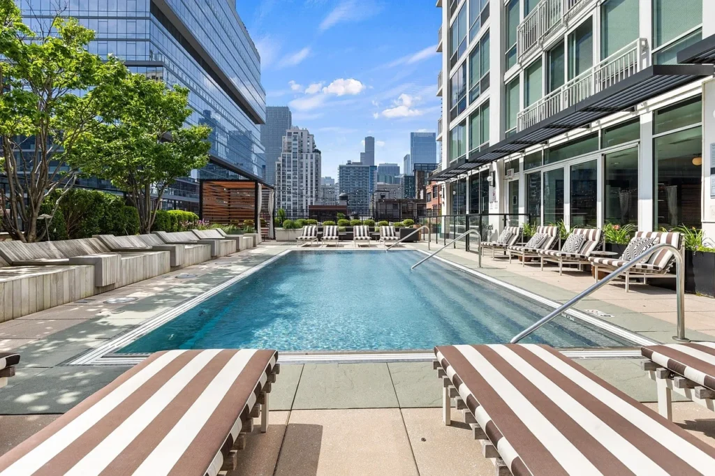 Rectangular outdoor pool surrounded by lounge chairs with striped cushions, next to a modern high-rise building, with city buildings in the background under a blue sky.