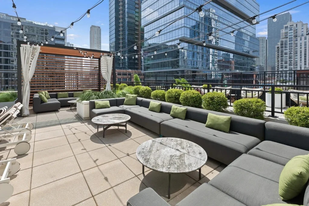 Rooftop patio with gray sofas, green cushions, round tables, potted plants, string lights, and city skyscrapers in the background under a blue sky.