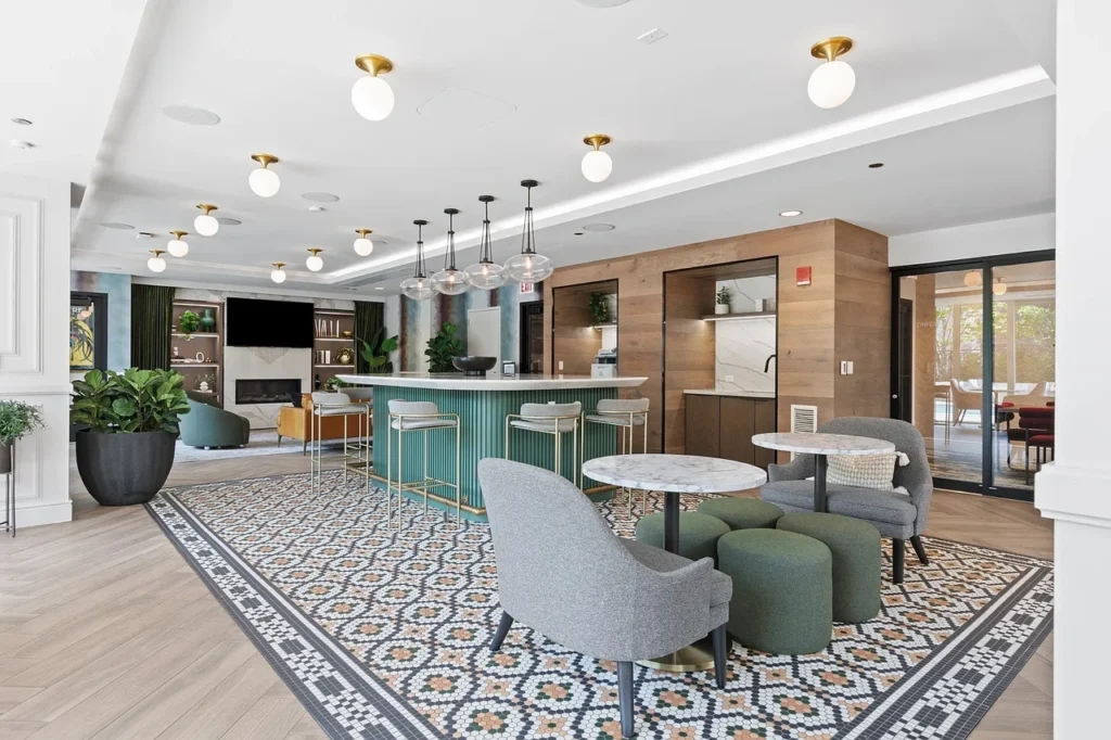 Modern lounge area with a green bar, round tables, gray chairs, patterned tile flooring, wood accents, and ceiling lights. Large windows and plants add natural light and greenery.