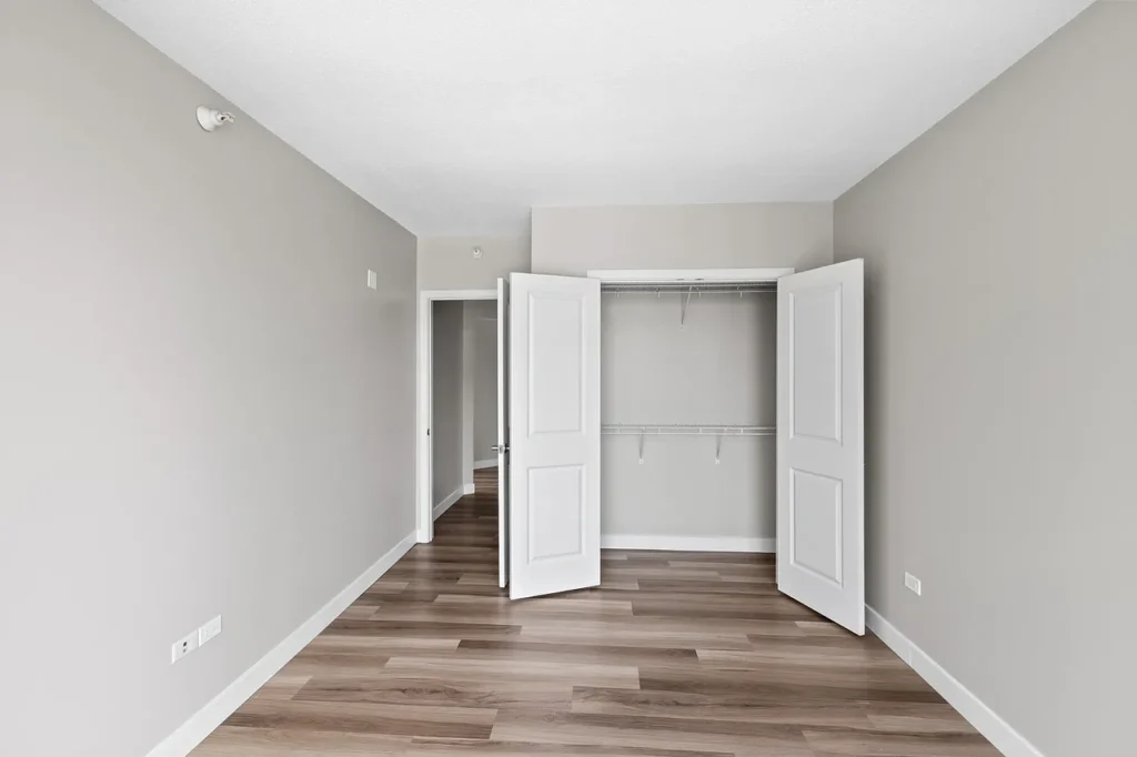 Empty room with light gray walls, open white double closet doors, wood flooring, and an open doorway leading to another room.