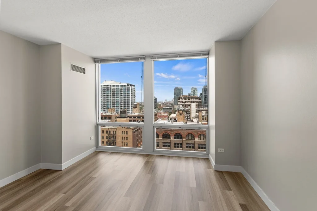 Unfurnished room with light wood flooring, beige walls, and large windows offering a cityscape view with several tall buildings under a blue sky.