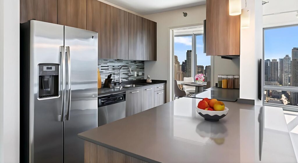 Modern kitchen with stainless steel appliances, wooden cabinets, a bowl of fruit on the counter, and city views visible through large windows.