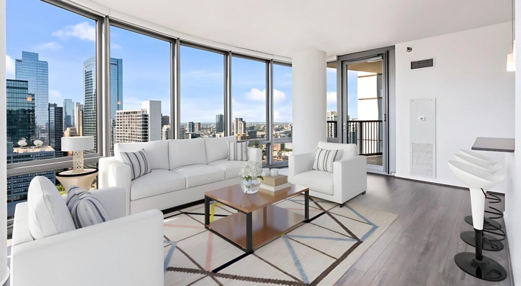 Modern living room with white sofas, glass coffee table, and large windows showing a city skyline; kitchen island with bar stools is visible on the right.