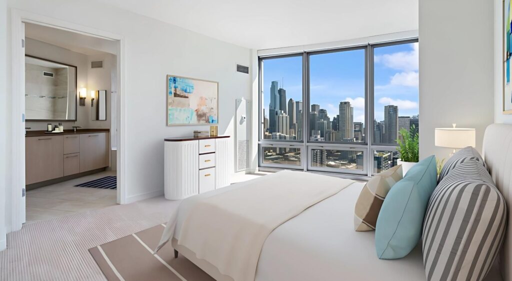Modern bedroom with a large bed, neutral decor, ensuite bathroom, and floor-to-ceiling windows overlooking a city skyline on a sunny day.