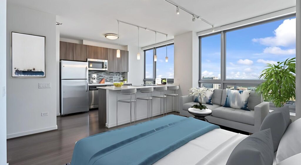 Modern studio apartment with a kitchen, breakfast bar, living area, large windows, and a bed in the foreground. Natural light fills the space, and a potted plant is by the window.