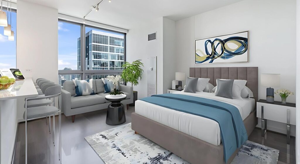 Modern bedroom with a large bed, blue and gray decor, a sofa, a breakfast bar with stools, and floor-to-ceiling windows overlooking city buildings.