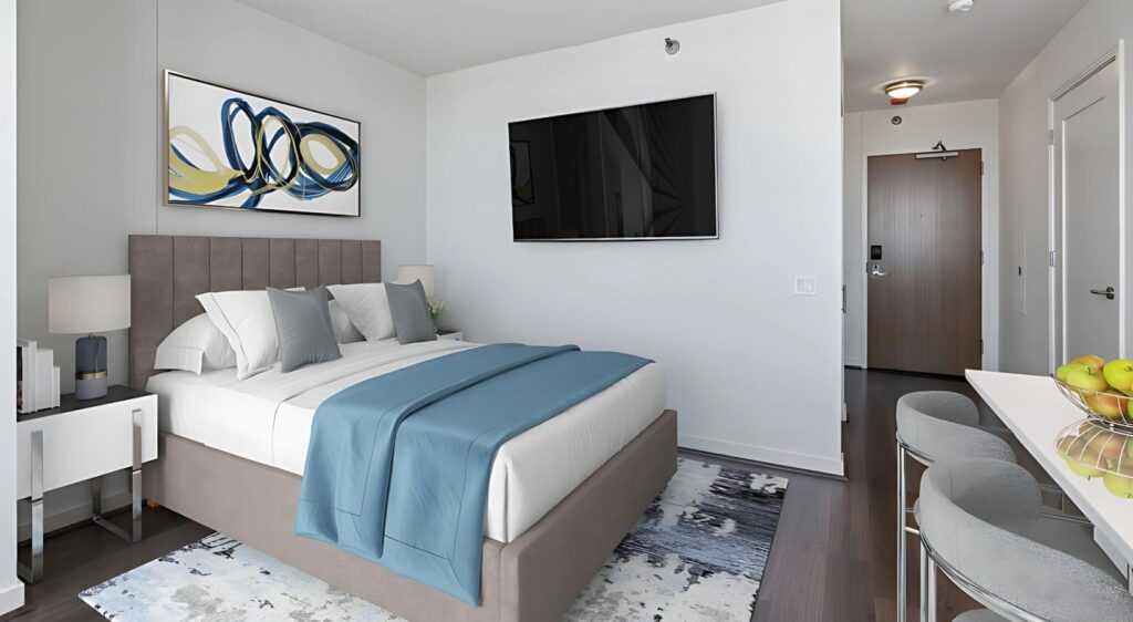 Modern hotel room with a large bed, blue and white bedding, wall-mounted TV, abstract artwork, and a small table with chairs; the entrance door is visible in the background.