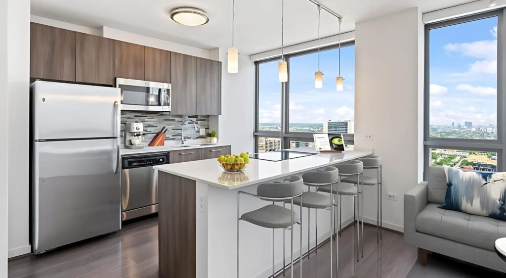 Modern kitchen with stainless steel appliances, white countertops, bar stools, pendant lights, and large windows offering a city skyline view.