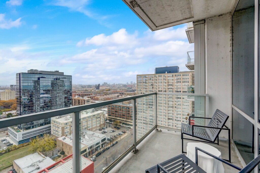 High-rise balcony with glass railing, two chairs, and a small table overlooking city buildings and a partly cloudy sky.