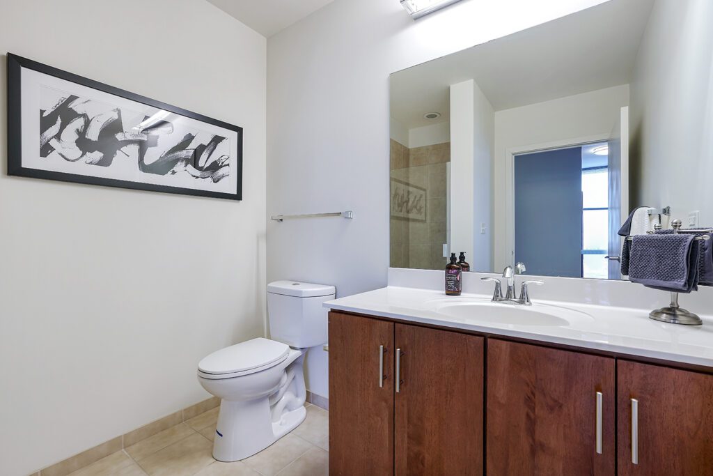 Modern bathroom with a large mirror, wooden vanity, white sink, toilet, wall art, and tiled floor. Navy towels and soap dispensers are on the countertop.