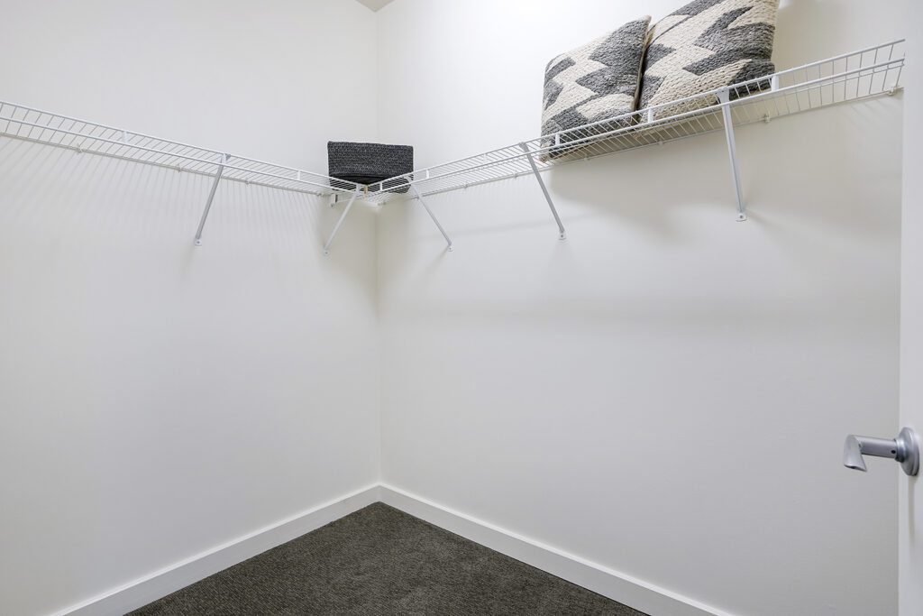 Empty walk-in closet with white walls, wire shelving, two patterned pillows, and a black basket on the top shelf.