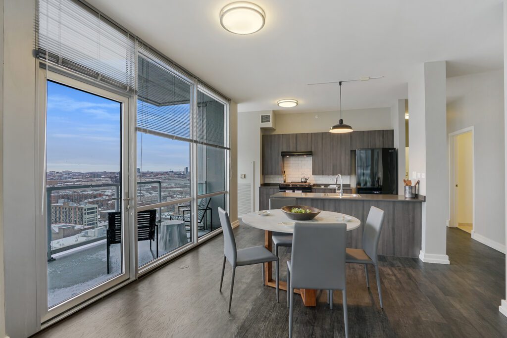Modern apartment kitchen and dining area with gray cabinets, round table, four chairs, large windows, and cityscape view.