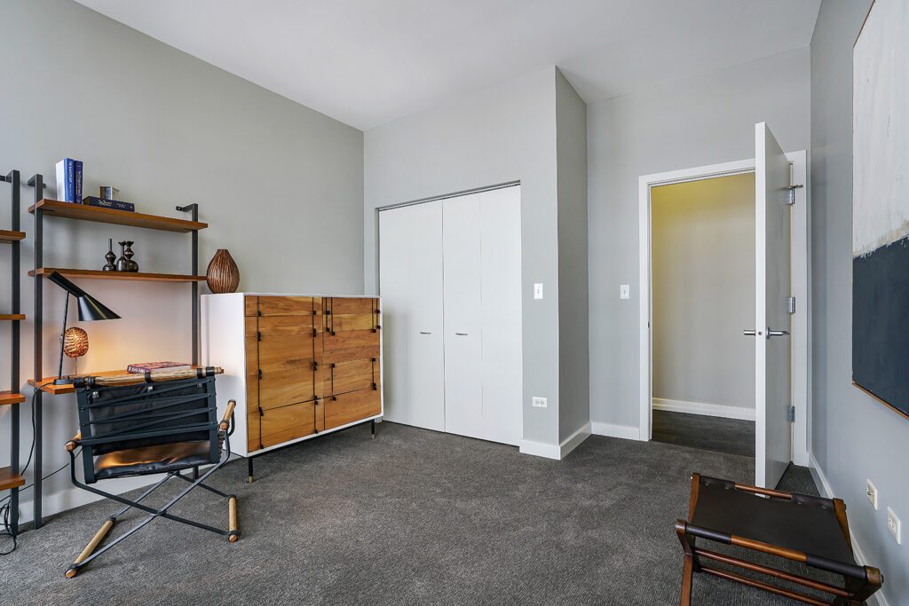 Modern home office with a wooden desk, black chair, wooden cabinet, wall shelving, grey carpet, and closed white closet doors.
