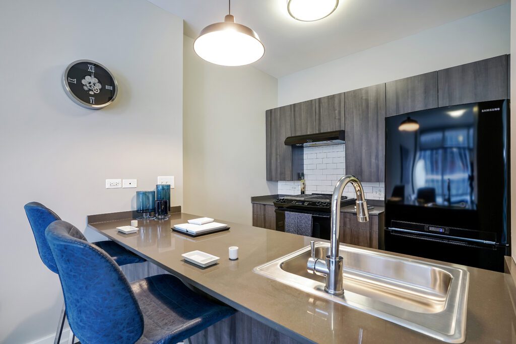 Modern kitchen with a dark countertop island, built-in sink, two blue chairs, stainless steel appliances, wall clock, and neutral-colored cabinets.