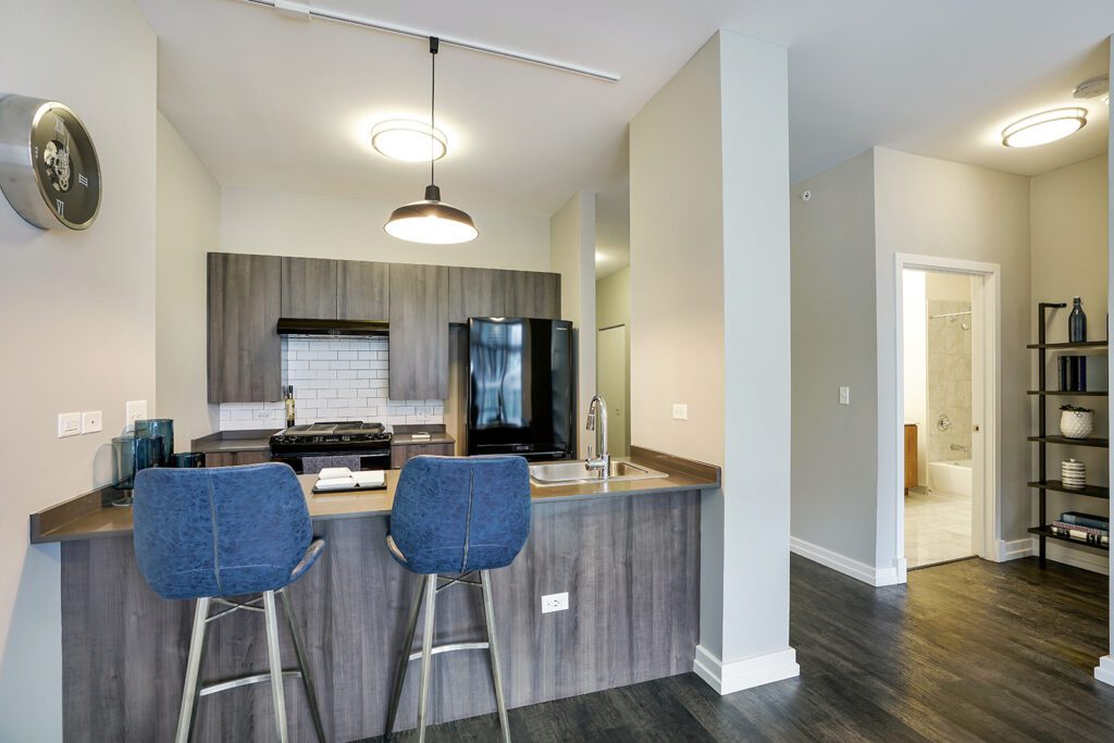 Modern kitchen with gray cabinets, black appliances, subway tile backsplash, two blue barstools at counter, open shelves, and view into a bathroom.