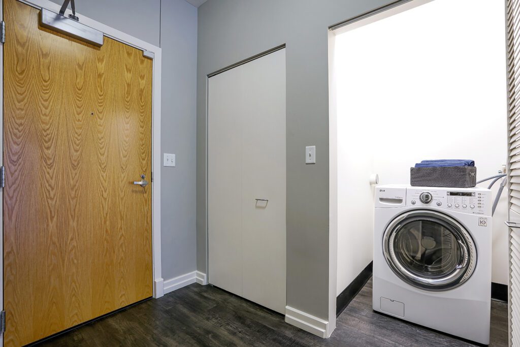 A front-loading washing machine with folded towels on top is in a small laundry nook next to a wooden door and a closed closet.