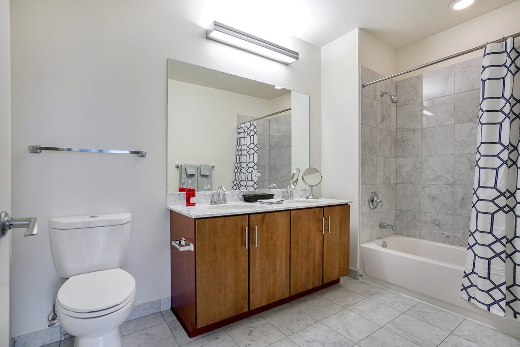 Modern bathroom with marble tile floor, wood vanity with double sinks, large mirror, toilet, and a bathtub with geometric-patterned shower curtain.