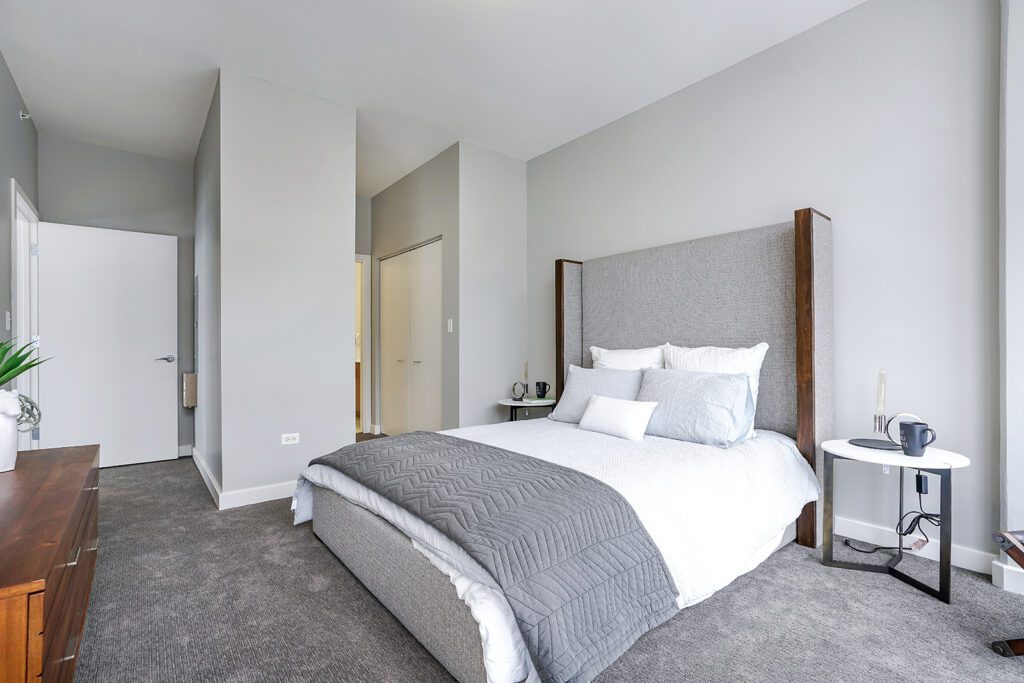 Modern bedroom with a large bed, gray headboard, white bedding, two side tables, gray carpet, and neutral walls. A door and closet are visible in the background.