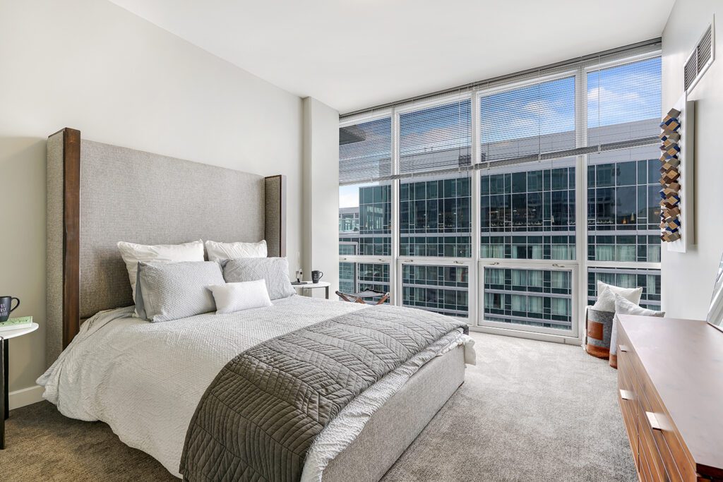 Modern bedroom with large windows, a gray upholstered bed, neutral bedding, and city building views outside. Minimalist decor includes side tables and a wooden dresser.