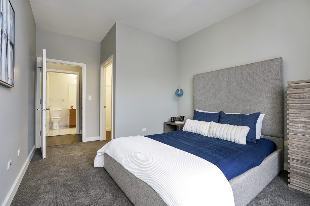 A modern bedroom with a gray upholstered bed, blue and white bedding, carpeted floor, and an open door revealing a bathroom in the background.