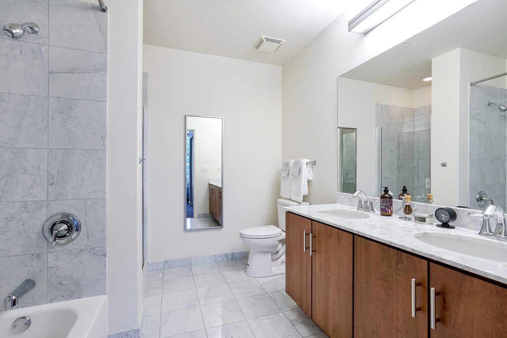 Modern bathroom with marble tile, double-sink vanity, large mirror, toilet, glass shower, bathtub, and towel rack on a light-colored wall.