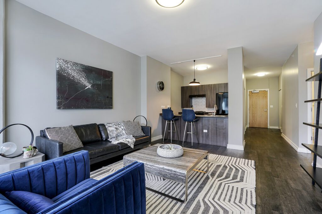 Modern open-concept living room with blue and black seating, geometric rug, abstract wall art, and a kitchen with bar stools in the background.