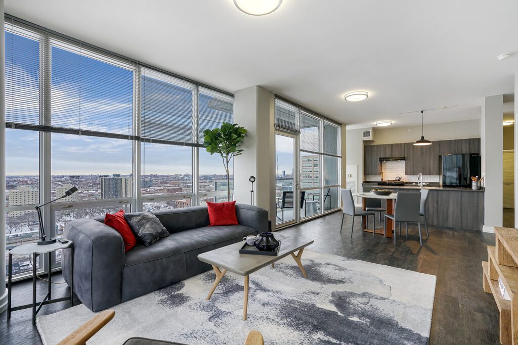 Modern apartment living room with large windows, gray sofa, kitchen area, dining table, and a city view in the background.