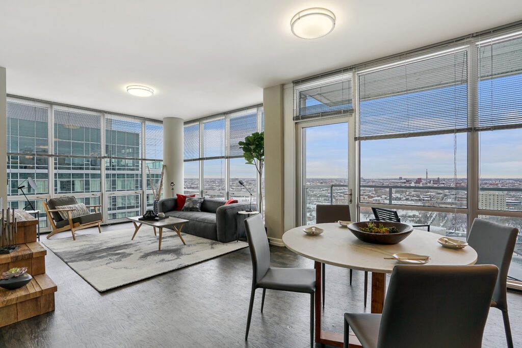 Modern apartment living room and dining area with large windows, city view, and contemporary furnishings including a round table, chairs, sofa, and potted plant.