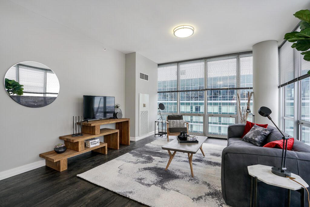 Modern living room with large windows, gray sofa, wooden furniture, wall-mounted TV, decorative items, and cityscape view in the background.