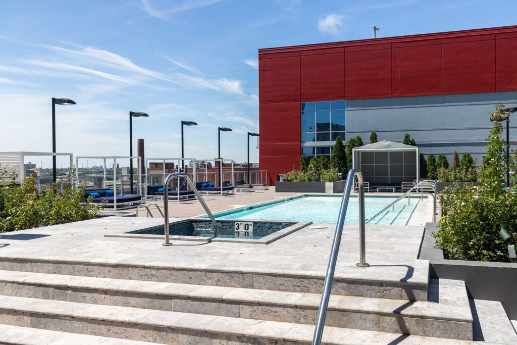 Rooftop pool and hot tub area with stairs, metal railings, and surrounding greenery beside a modern red and gray building under a clear sky.