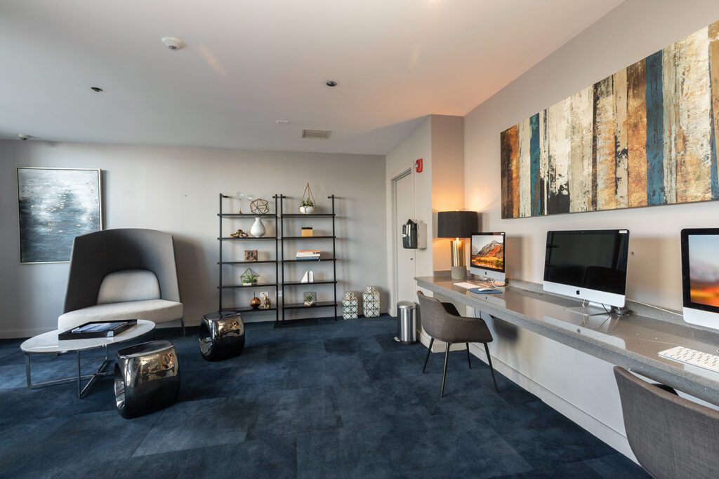 Modern office lounge with blue carpet, open shelving, abstract wall art, a lounge chair, and a long desk with two computers and chairs.