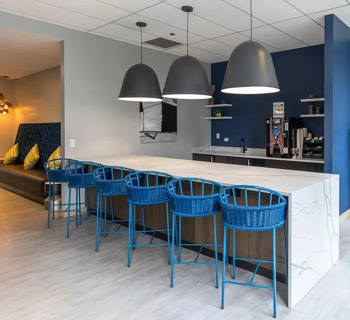 Modern kitchen area with a marble island, five blue bar stools, pendant lights overhead, a coffee machine, and built-in seating with yellow cushions along the wall.