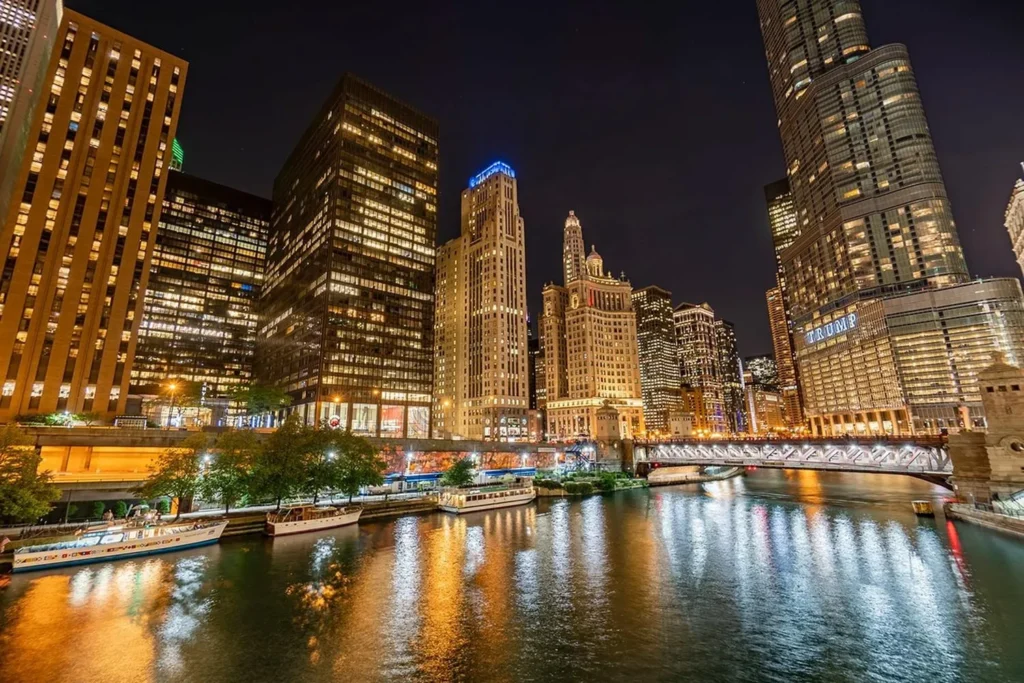 Night view of a city skyline with illuminated buildings, a lit bridge, and boats docked along a river reflecting the city lights.