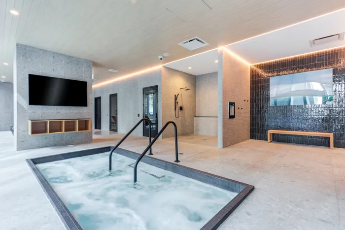 Modern indoor spa room featuring a rectangular hot tub with bubbling water, a wall-mounted TV, a shower area, and a bench next to a tiled accent wall.