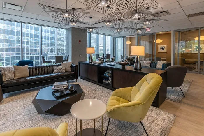 Modern office lounge with large windows, stylish light fixtures, yellow chairs, a black sofa, and a central coffee table on a gray rug, with city buildings visible outside.