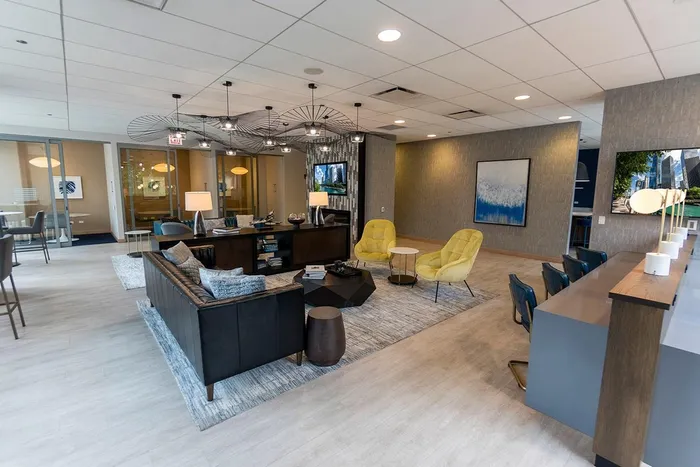Modern lounge area with sofas, yellow chairs, bar seating, abstract wall art, and large ceiling light fixtures in a spacious, well-lit room.