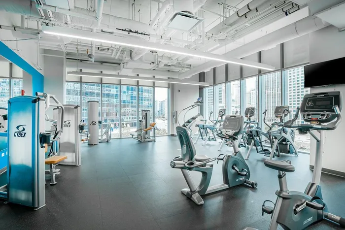 Modern gym with various exercise machines including treadmills, stationary bikes, and weight equipment, featuring large windows and city views.