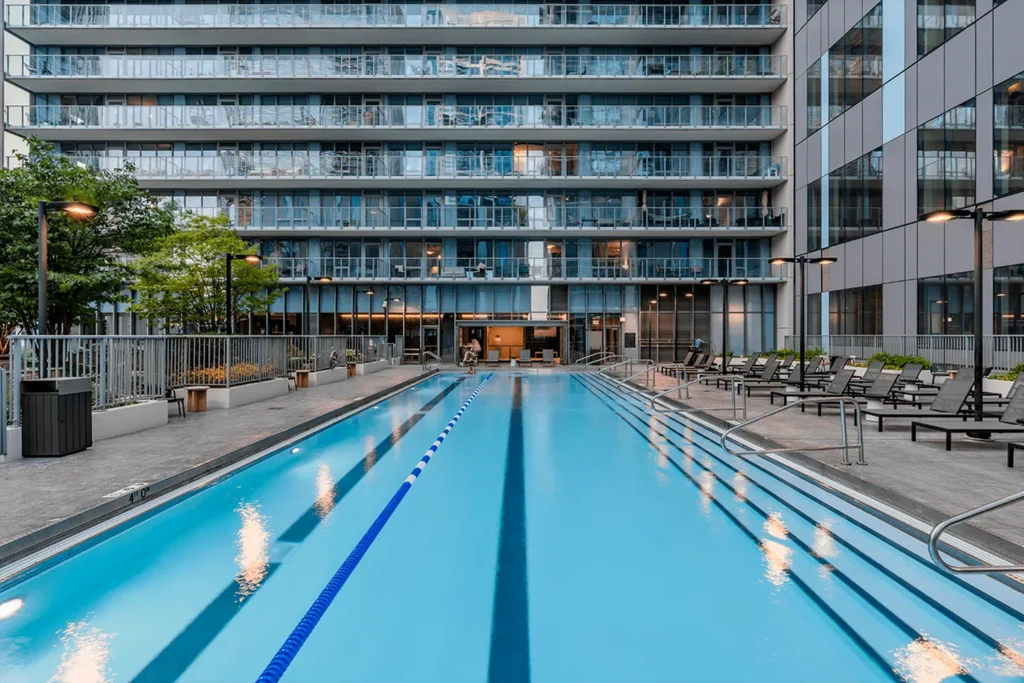 A modern outdoor lap pool with a blue lane divider, surrounded by lounge chairs and tall glass apartment buildings.