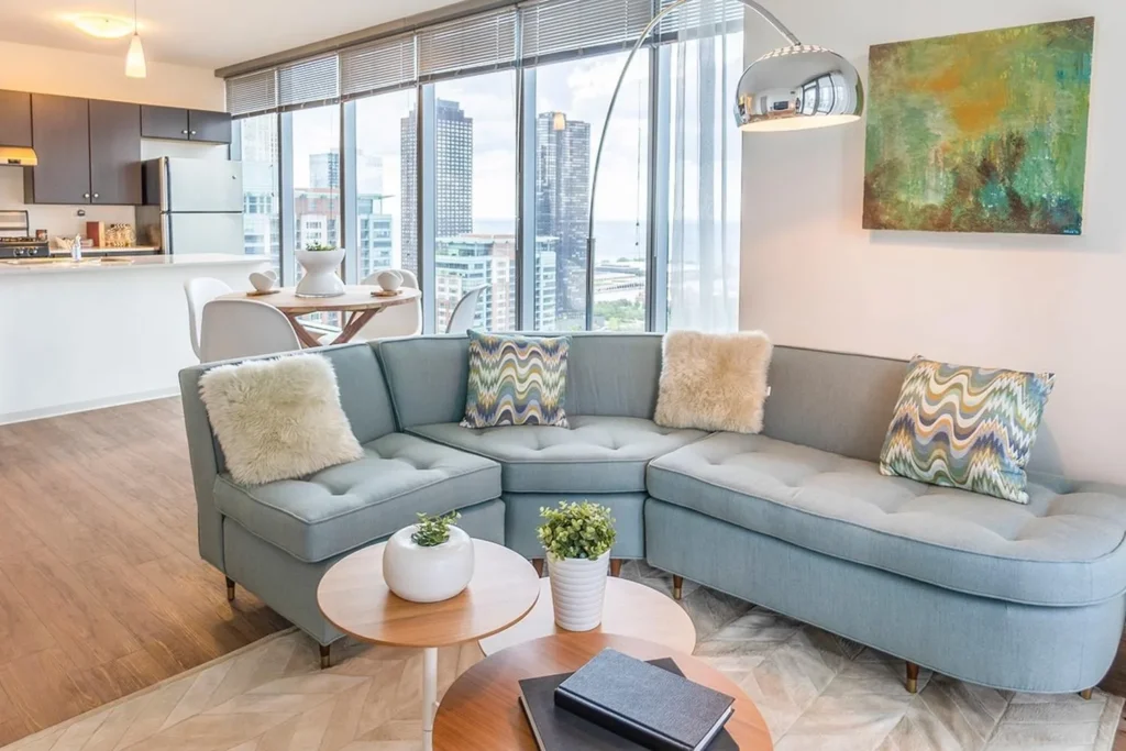 Modern living room with a light blue sectional sofa, patterned pillows, round wooden coffee tables, plants, large windows, and a view of city skyscrapers. Kitchen and dining area in background.