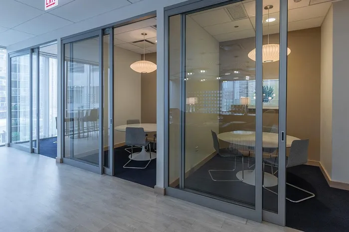 Three glass-walled office meeting rooms with round tables, chairs, and pendant lights, located off a hallway with light wood flooring and large windows.