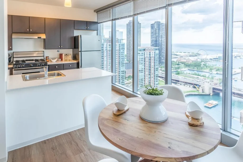 Modern kitchen and dining area with wood table, white chairs, and large windows overlooking city buildings and a waterfront.