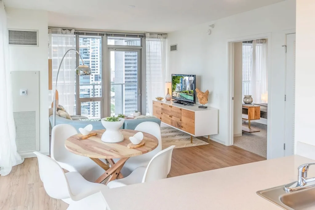 Modern open-plan living space with a round dining table, white chairs, a TV, and large windows overlooking city buildings. A bedroom is visible through an open door.